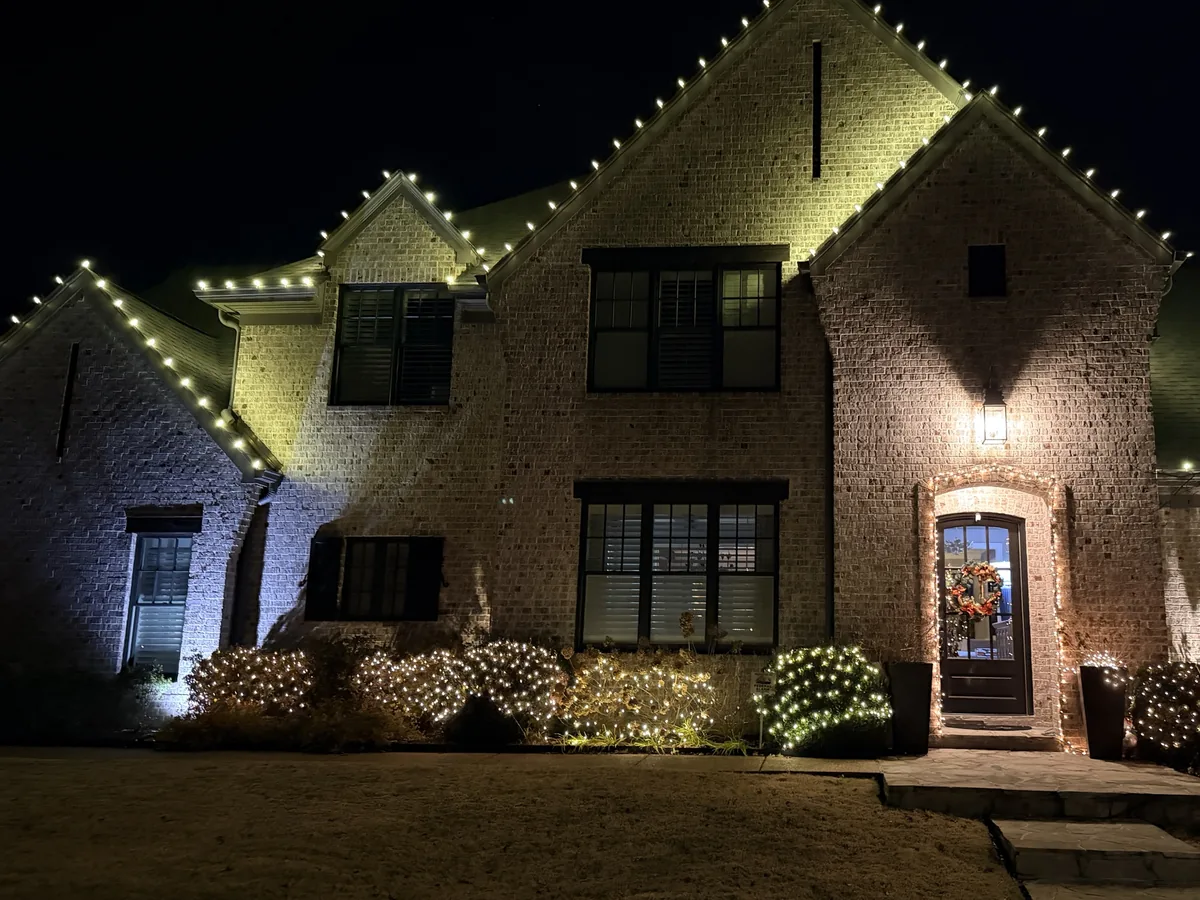 Two-story brick home with warm white roofline lights, lit shrubs, and a lit front doorway